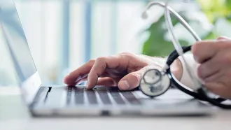 Doctor's hand typing on a laptop, stethoscope in the other hand.